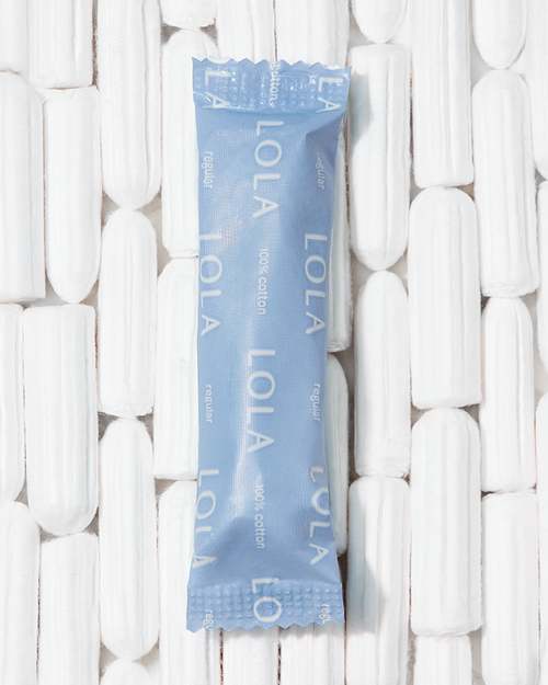 A close up of a Lola tampon in a package with unpacked tampons arranged in a grid in the background